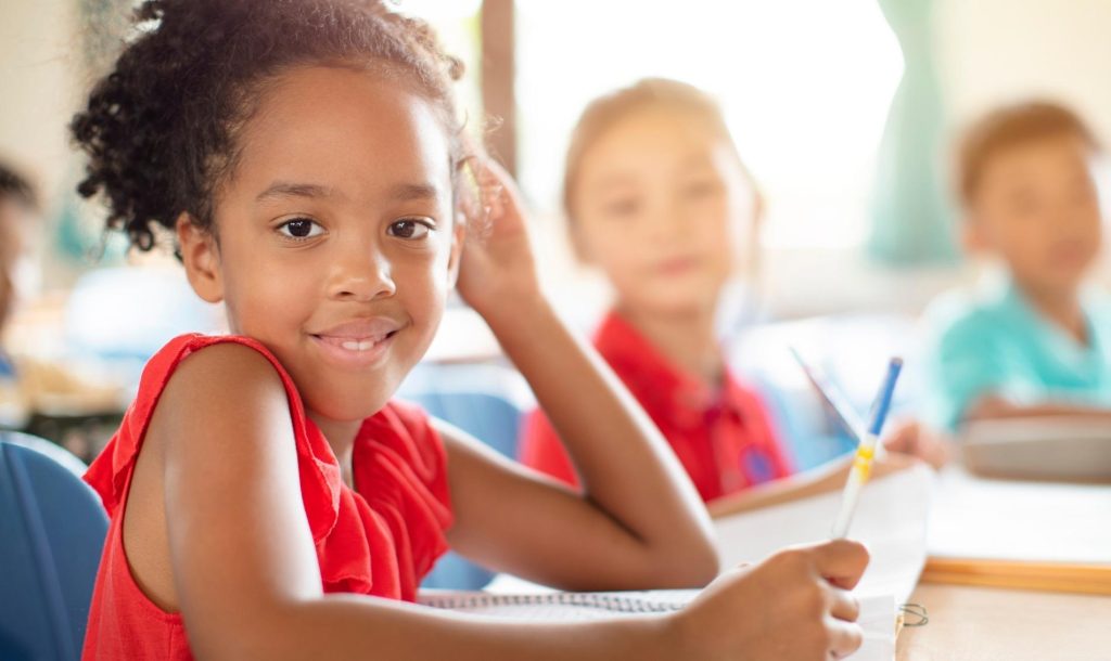 Happy elementary student at desk with two other students in the background