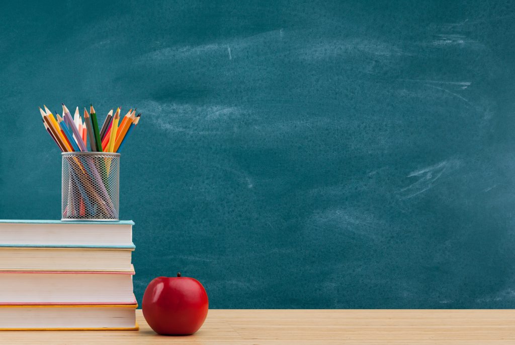 A cup of colored pencils sits on a stack of four text books, next to a red apple. A green chalkboard is in the background.