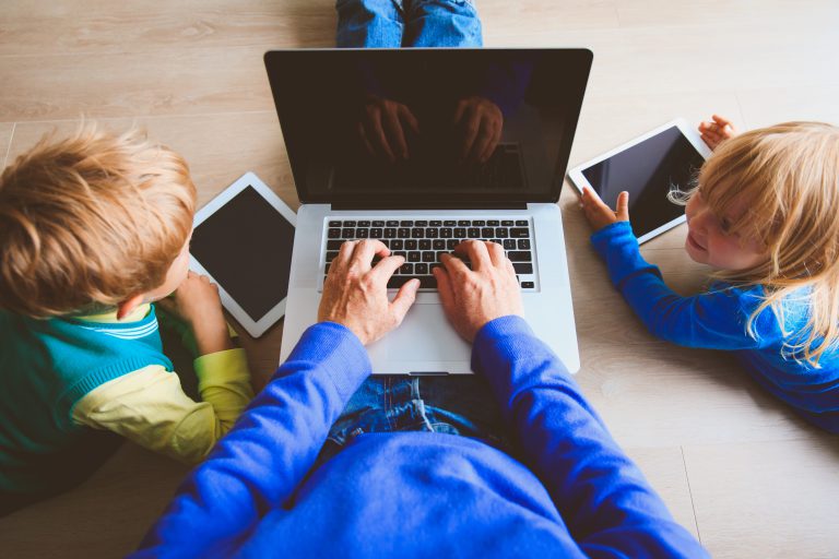 Two children with tablets watch as an adult types on a laptop