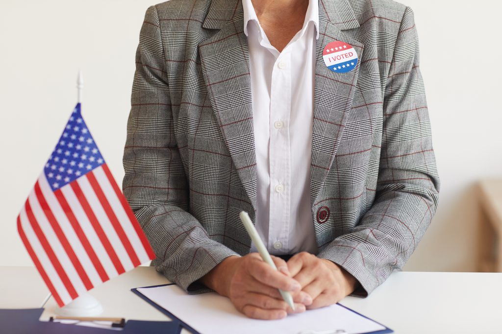 Person wearing a blazer holds a pen over a clip board. They wear an "I Voted" sticker