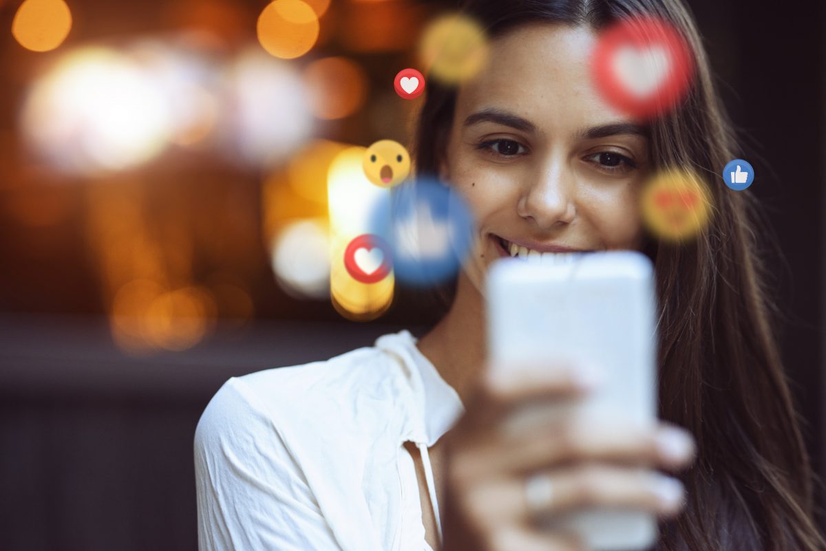 A woman smiles while holding a cellphone. Around her face are reaction emojis