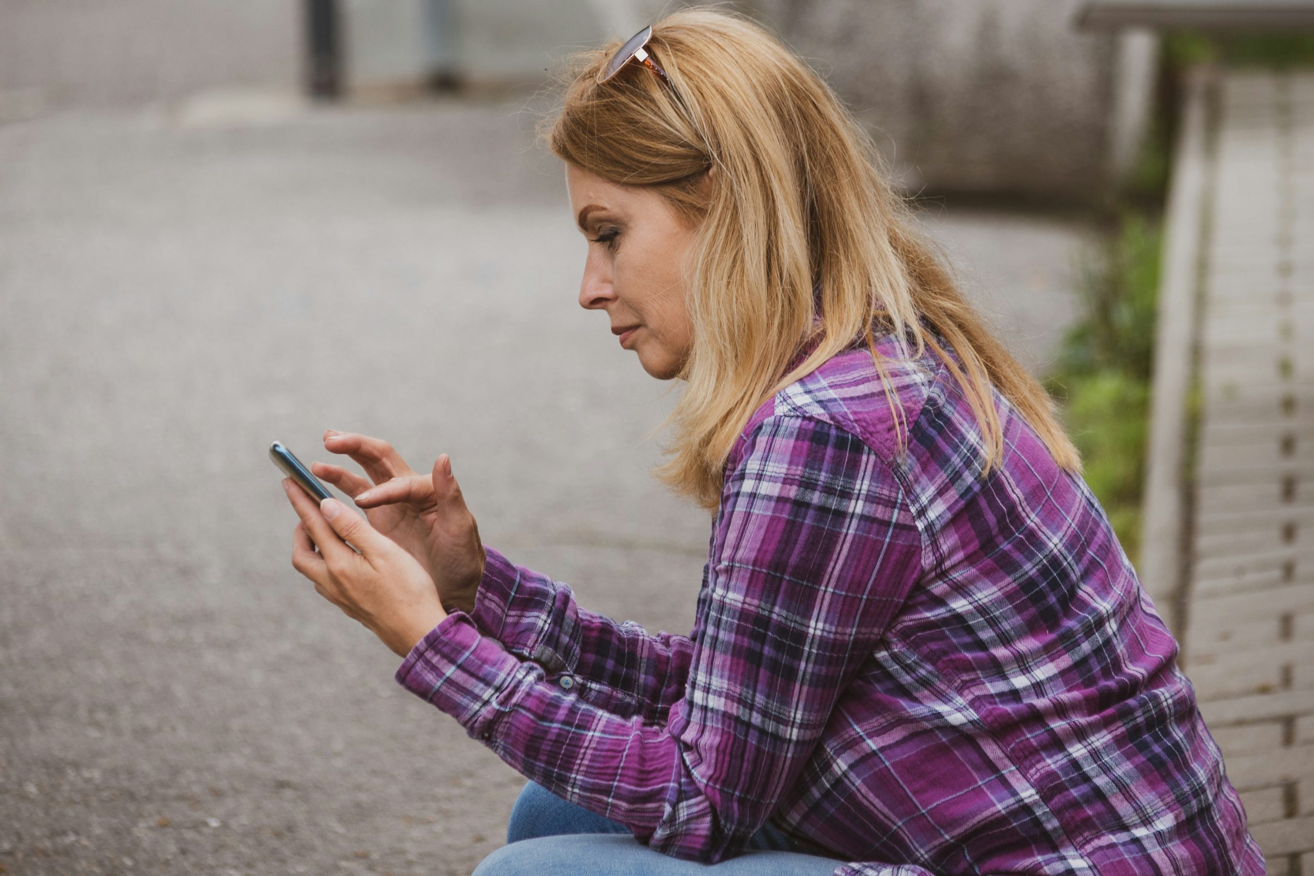 Woman with blonde hair and a plaid purple shirt sits on the curb of a quiet street while looking at her smartphone.